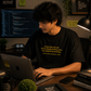 A man wearing a black graphic t-shirt with a yellow quote sits at a desk working on a laptop in a cozy coding room environment.